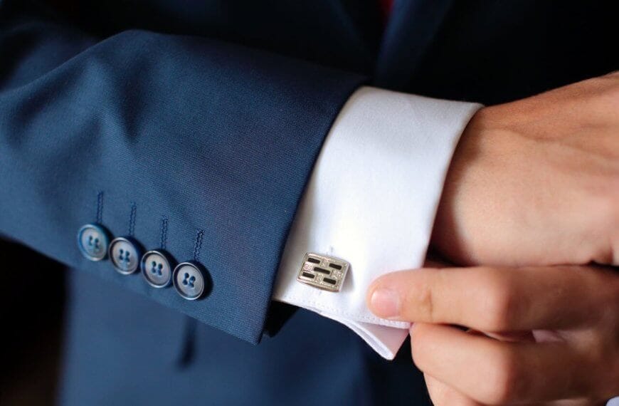 A person expertly adjusts a metallic, rectangular cufflink on a fitted white dress shirt. The tailored sleeve, part of a dark blue suit with four blue buttons, exudes elegance. The setting is shadowed and elusive, reminiscent of Metro Manila's sophisticated style.