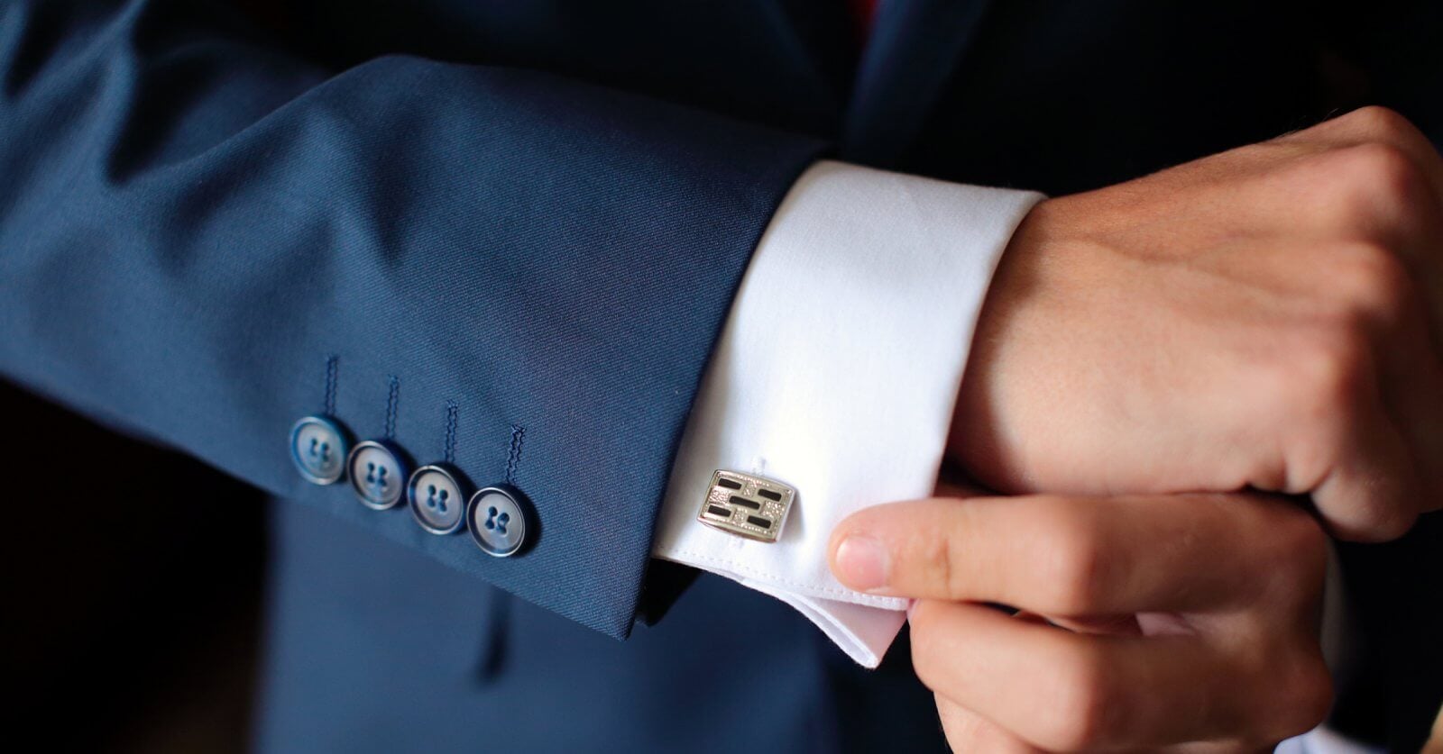 A person expertly adjusts a metallic, rectangular cufflink on a fitted white dress shirt. The tailored sleeve, part of a dark blue suit with four blue buttons, exudes elegance. The setting is shadowed and elusive, reminiscent of Metro Manila's sophisticated style.