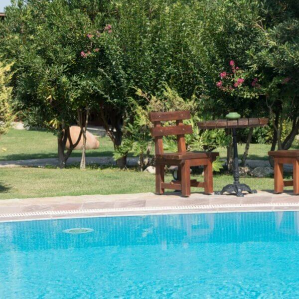 A serene outdoor scene with a clear blue swimming pool in the foreground, reminiscent of Calamba's famous hot springs. In the background, two wooden chairs and a small round table rest on a lush lawn in Laguna. Surrounding bushes and trees add nature's touch and privacy to the setting.