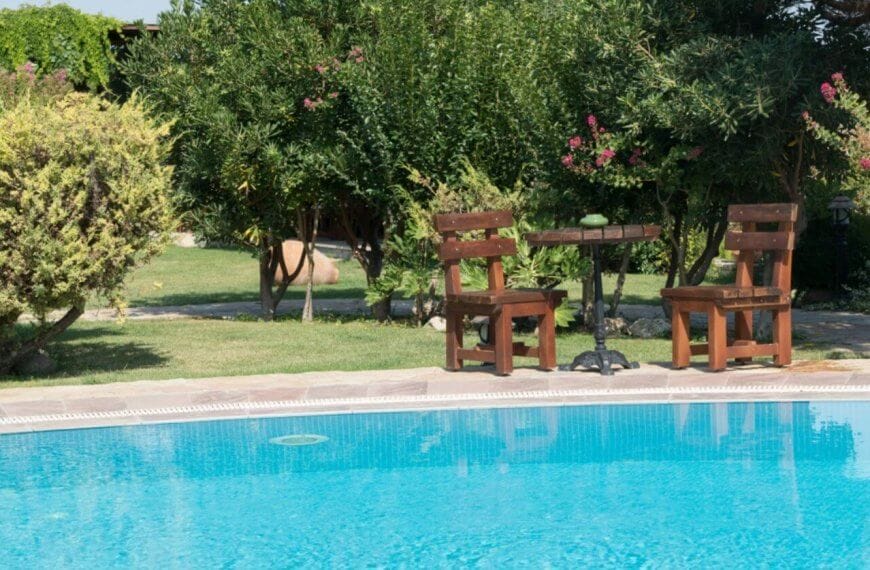 A serene outdoor scene with a clear blue swimming pool in the foreground, reminiscent of Calamba's famous hot springs. In the background, two wooden chairs and a small round table rest on a lush lawn in Laguna. Surrounding bushes and trees add nature's touch and privacy to the setting.