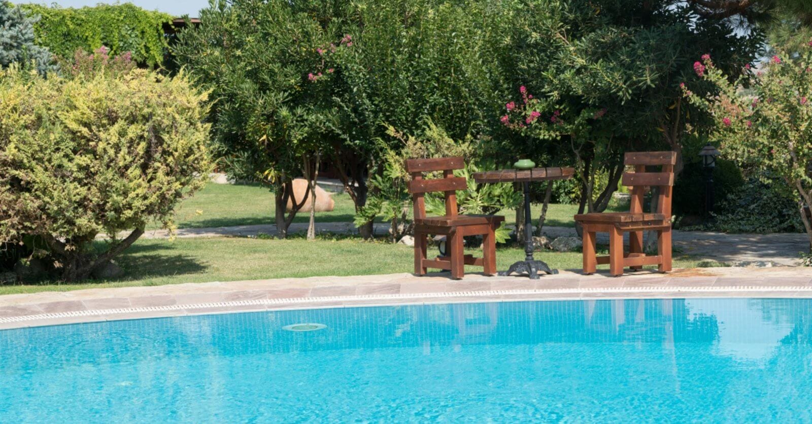 A serene outdoor scene with a clear blue swimming pool in the foreground, reminiscent of Calamba's famous hot springs. In the background, two wooden chairs and a small round table rest on a lush lawn in Laguna. Surrounding bushes and trees add nature's touch and privacy to the setting.