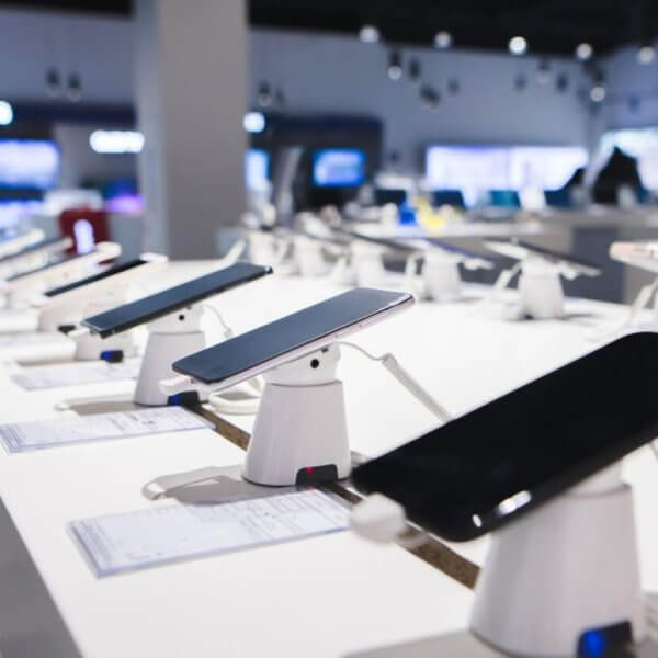 Smartphones are displayed on a white counter in a DITO Experience Store, each mounted on a stand with security features. The store, located in the Philippines, has bright lighting and a blurred background showcasing more electronic devices, with price tags accompanying each phone.