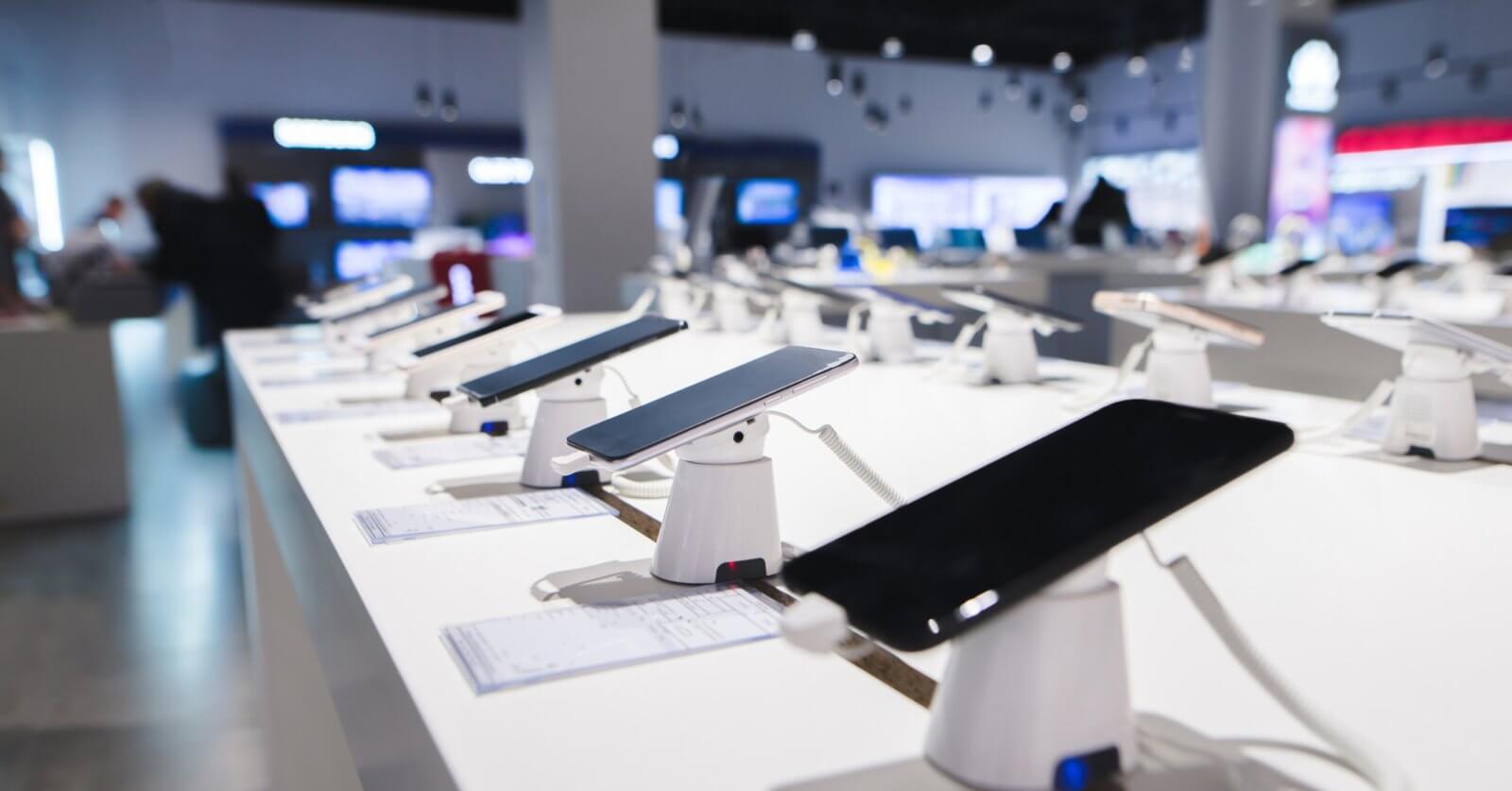 Smartphones are displayed on a white counter in a DITO Experience Store, each mounted on a stand with security features. The store, located in the Philippines, has bright lighting and a blurred background showcasing more electronic devices, with price tags accompanying each phone.