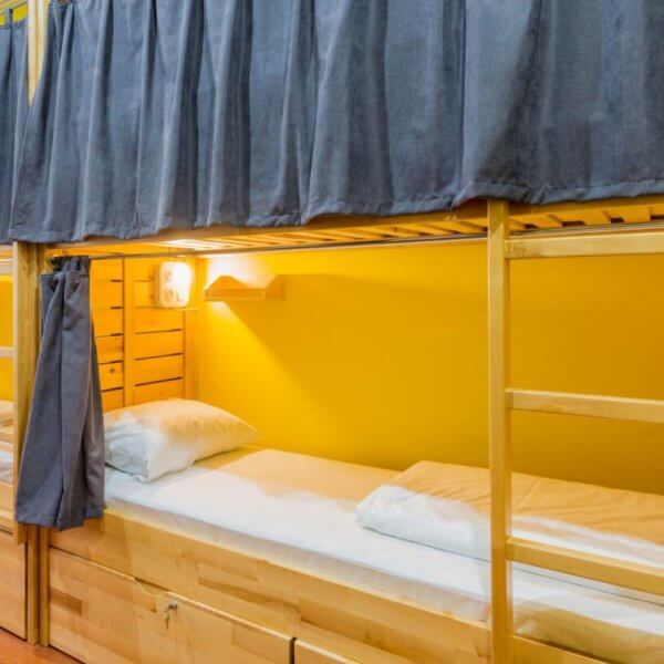 Cozy bunk beds with wooden frames, each adorned with navy curtains, pillows, and lamps, rest against a yellow wall in the best dormitory in Metro Manila.