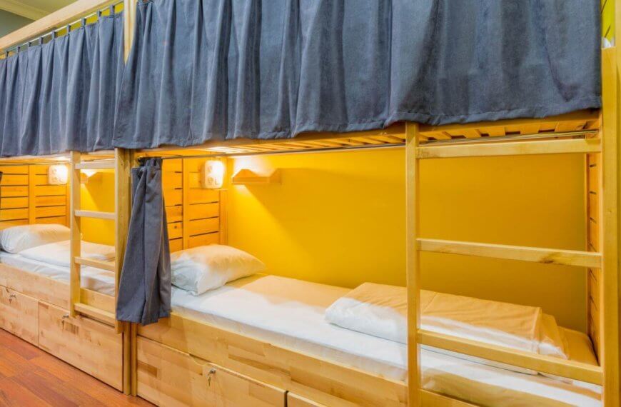 Cozy bunk beds with wooden frames, each adorned with navy curtains, pillows, and lamps, rest against a yellow wall in the best dormitory in Metro Manila.