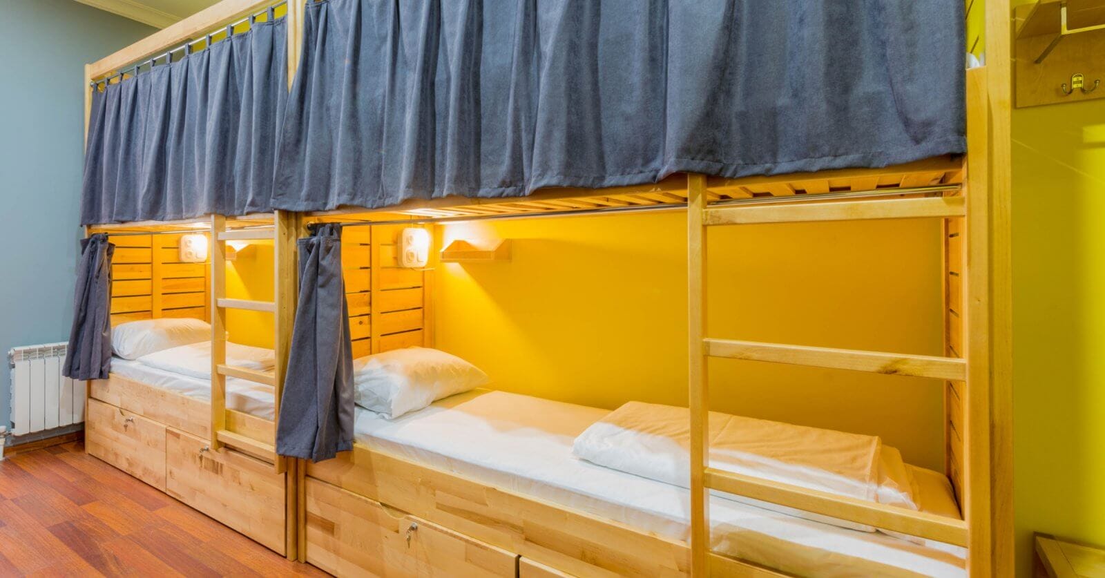 Cozy bunk beds with wooden frames, each adorned with navy curtains, pillows, and lamps, rest against a yellow wall in the best dormitory in Metro Manila.