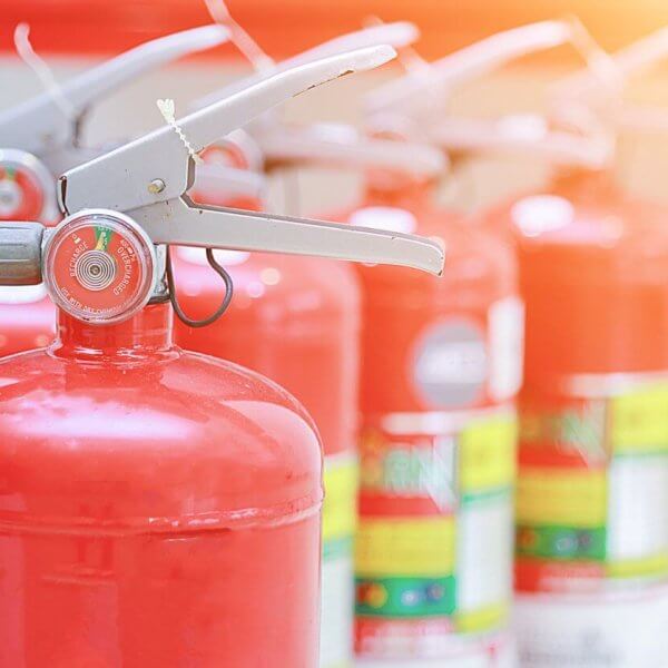 Close-up of red fire extinguishers, supplied by top Fire Extinguisher Suppliers in Metro Manila, standing in a row. The foreground extinguisher is in focus, its nozzle and pressure gauge clear, while others blur into the background. The scene is brightly lit with a warm glow in the top right corner.
