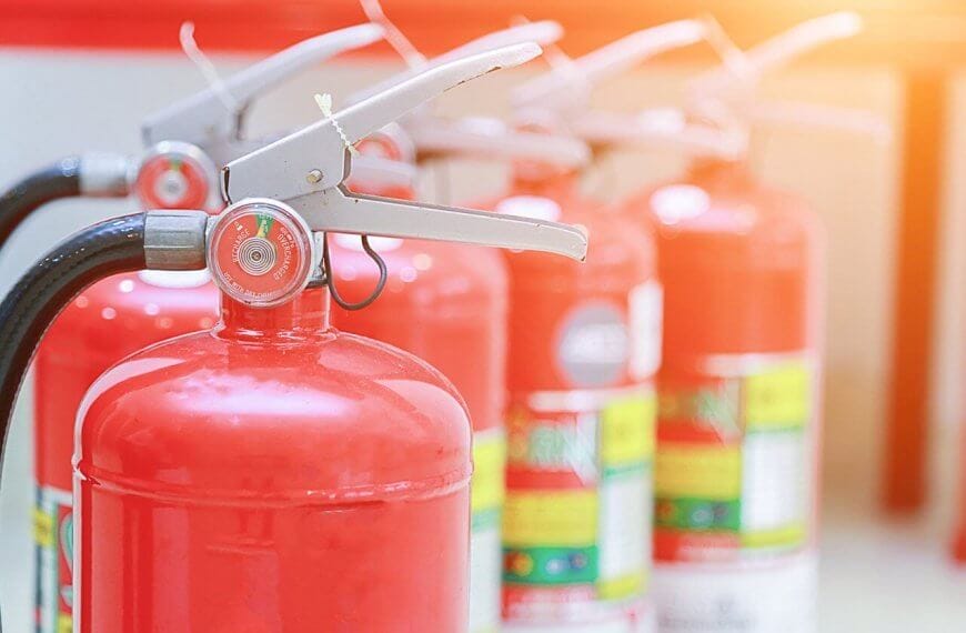 Close-up of red fire extinguishers, supplied by top Fire Extinguisher Suppliers in Metro Manila, standing in a row. The foreground extinguisher is in focus, its nozzle and pressure gauge clear, while others blur into the background. The scene is brightly lit with a warm glow in the top right corner.