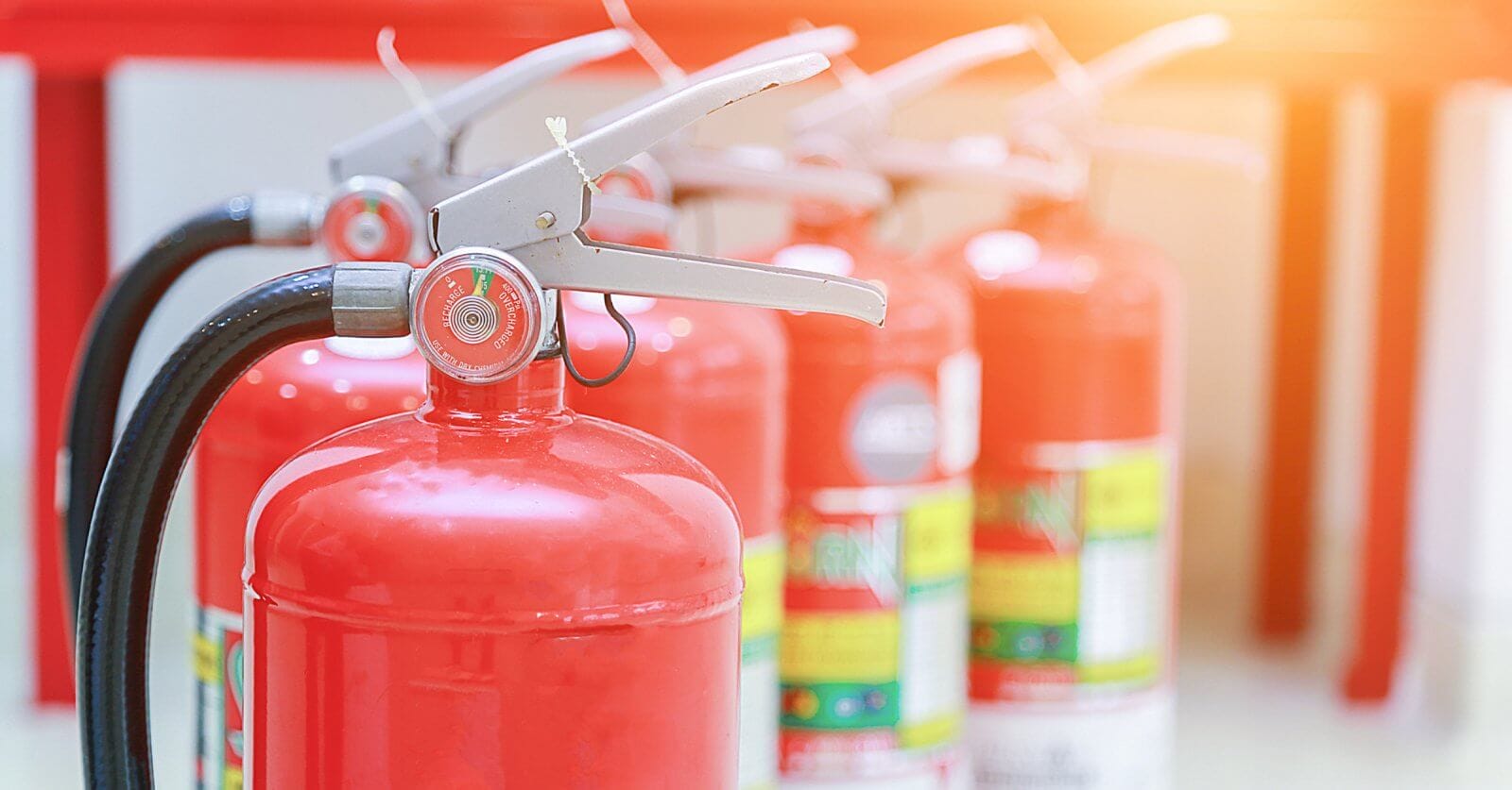 Close-up of red fire extinguishers, supplied by top Fire Extinguisher Suppliers in Metro Manila, standing in a row. The foreground extinguisher is in focus, its nozzle and pressure gauge clear, while others blur into the background. The scene is brightly lit with a warm glow in the top right corner.