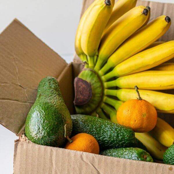 A cardboard box from a food distributor in Metro Manila is filled with bananas, avocados, and oranges. A plastic cup containing red and yellow cherry tomatoes is also visible. An apple rests at the front of the open box, showcasing the colorful and fresh produce against a neutral background.