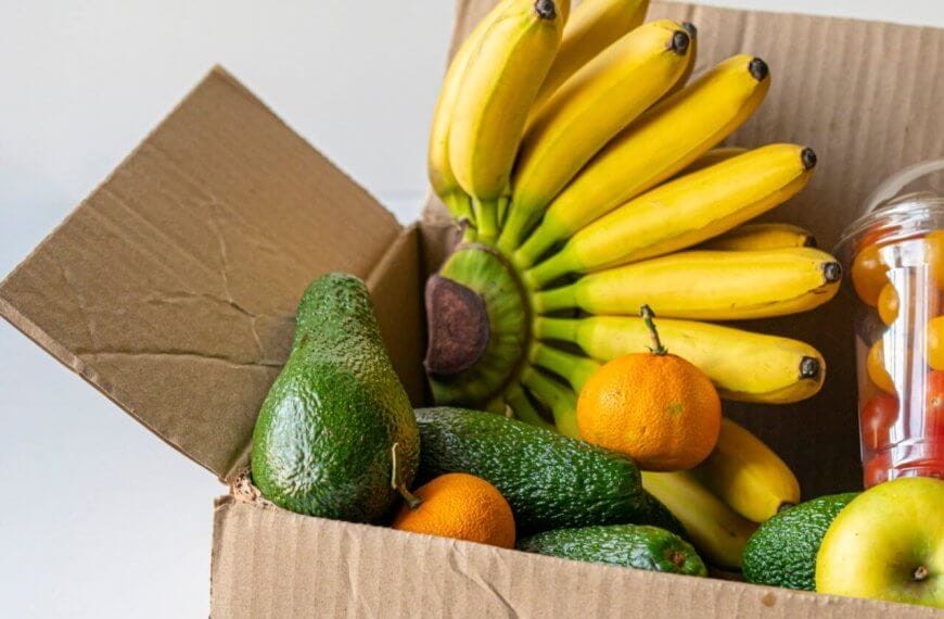 A cardboard box from a food distributor in Metro Manila is filled with bananas, avocados, and oranges. A plastic cup containing red and yellow cherry tomatoes is also visible. An apple rests at the front of the open box, showcasing the colorful and fresh produce against a neutral background.
