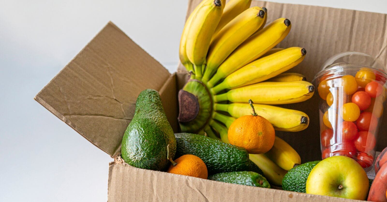 A cardboard box from a food distributor in Metro Manila is filled with bananas, avocados, and oranges. A plastic cup containing red and yellow cherry tomatoes is also visible. An apple rests at the front of the open box, showcasing the colorful and fresh produce against a neutral background.