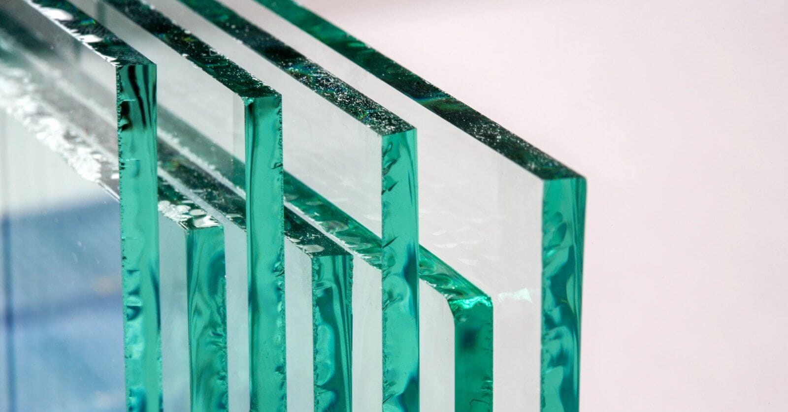 Close-up of four rectangular glass panes, supplied by leading glass suppliers in Metro Manila, standing upright and lined up horizontally against a neutral background. The edges show a green tint, with surfaces reflecting light to create a layered, transparent effect that highlights the clarity and texture.