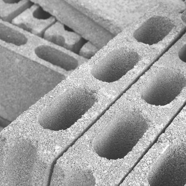 A close-up of stacked concrete cinder blocks, sourced from leading hollow block suppliers in Metro Manila, forms a monochrome setting. These blocks, with three elongated oval holes and rough textures, are arranged diagonally. Some are fully visible while others are partially obscured, crafting a repetitive geometric pattern.