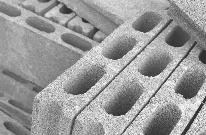 A close-up of stacked concrete cinder blocks, sourced from leading hollow block suppliers in Metro Manila, forms a monochrome setting. These blocks, with three elongated oval holes and rough textures, are arranged diagonally. Some are fully visible while others are partially obscured, crafting a repetitive geometric pattern.