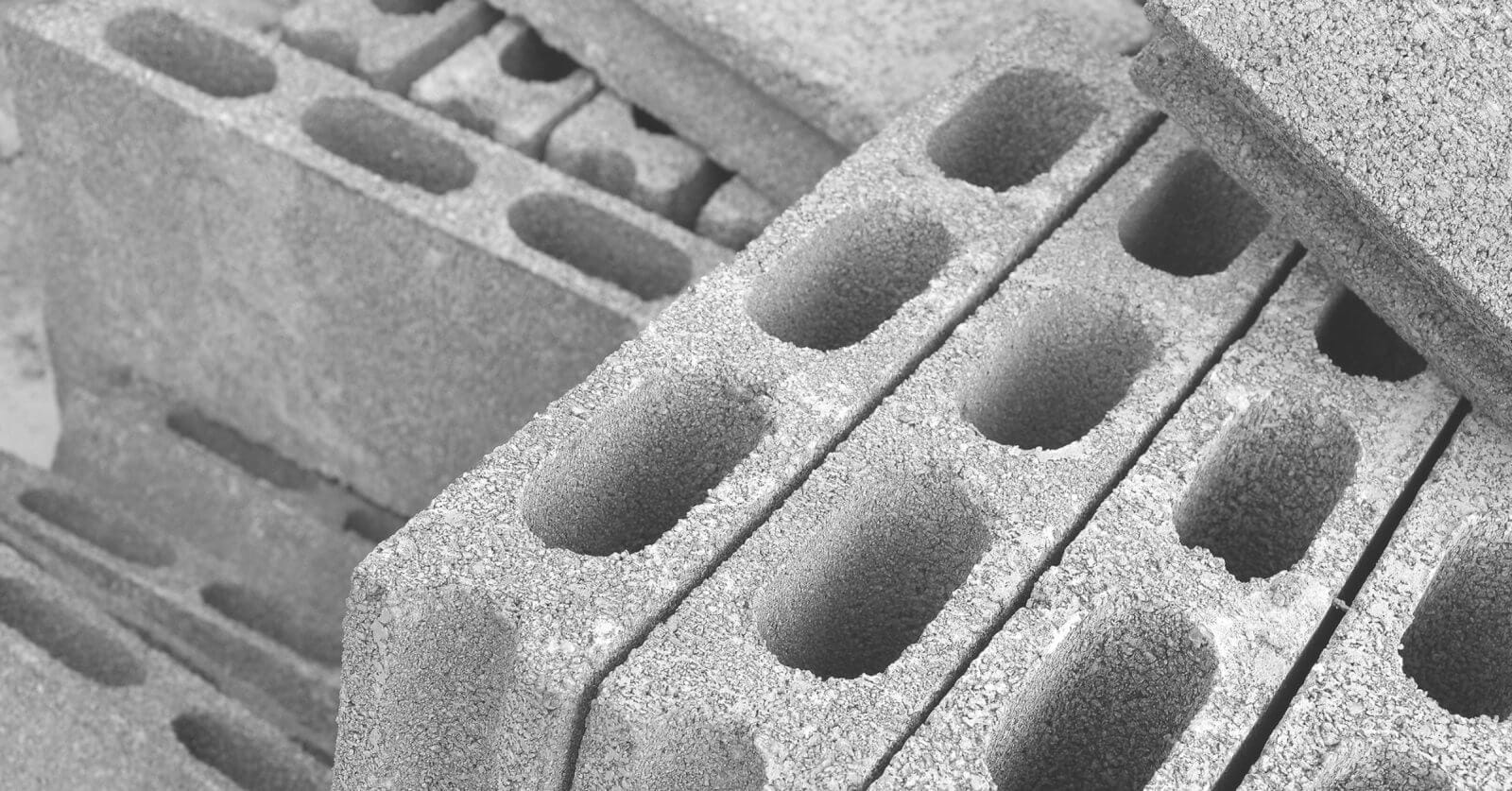A close-up of stacked concrete cinder blocks, sourced from leading hollow block suppliers in Metro Manila, forms a monochrome setting. These blocks, with three elongated oval holes and rough textures, are arranged diagonally. Some are fully visible while others are partially obscured, crafting a repetitive geometric pattern.