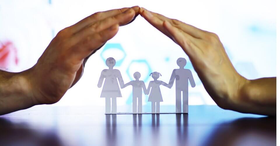 Hands forming a roof over paper cutout family figures on a table, symbolizing the meaningful impact of protection and security.
