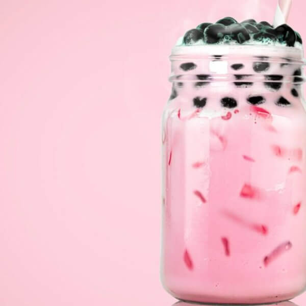 A clear glass mason jar filled with pink bubble tea from top Milktea suppliers, featuring black tapioca pearls on top. The drink contains ice cubes and is set against a solid pink background. A straw is inserted into the jar, evoking the refreshing, sweet flavors cherished in Metro Manila.