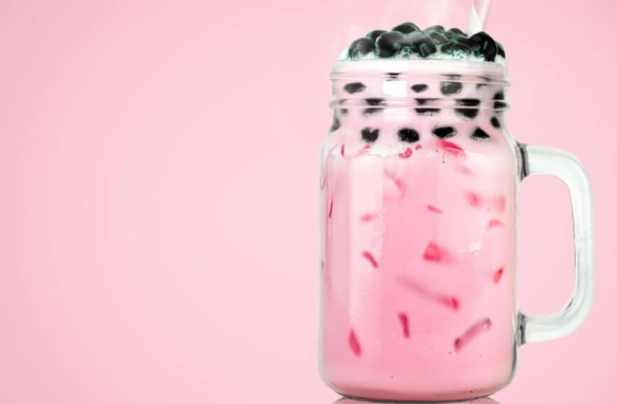A clear glass mason jar filled with pink bubble tea from top Milktea suppliers, featuring black tapioca pearls on top. The drink contains ice cubes and is set against a solid pink background. A straw is inserted into the jar, evoking the refreshing, sweet flavors cherished in Metro Manila.