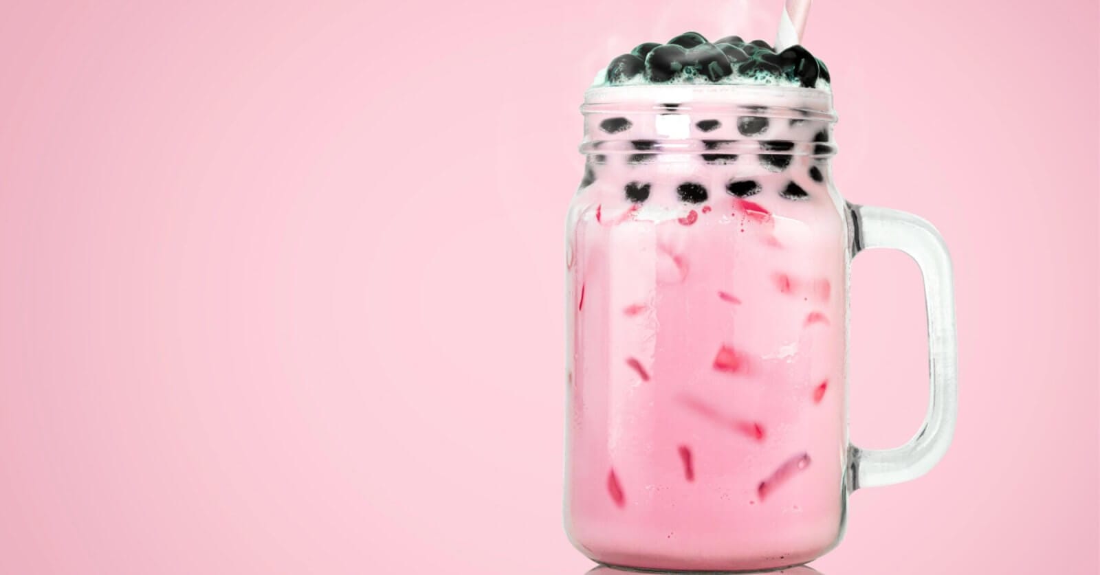 A clear glass mason jar filled with pink bubble tea from top Milktea suppliers, featuring black tapioca pearls on top. The drink contains ice cubes and is set against a solid pink background. A straw is inserted into the jar, evoking the refreshing, sweet flavors cherished in Metro Manila.