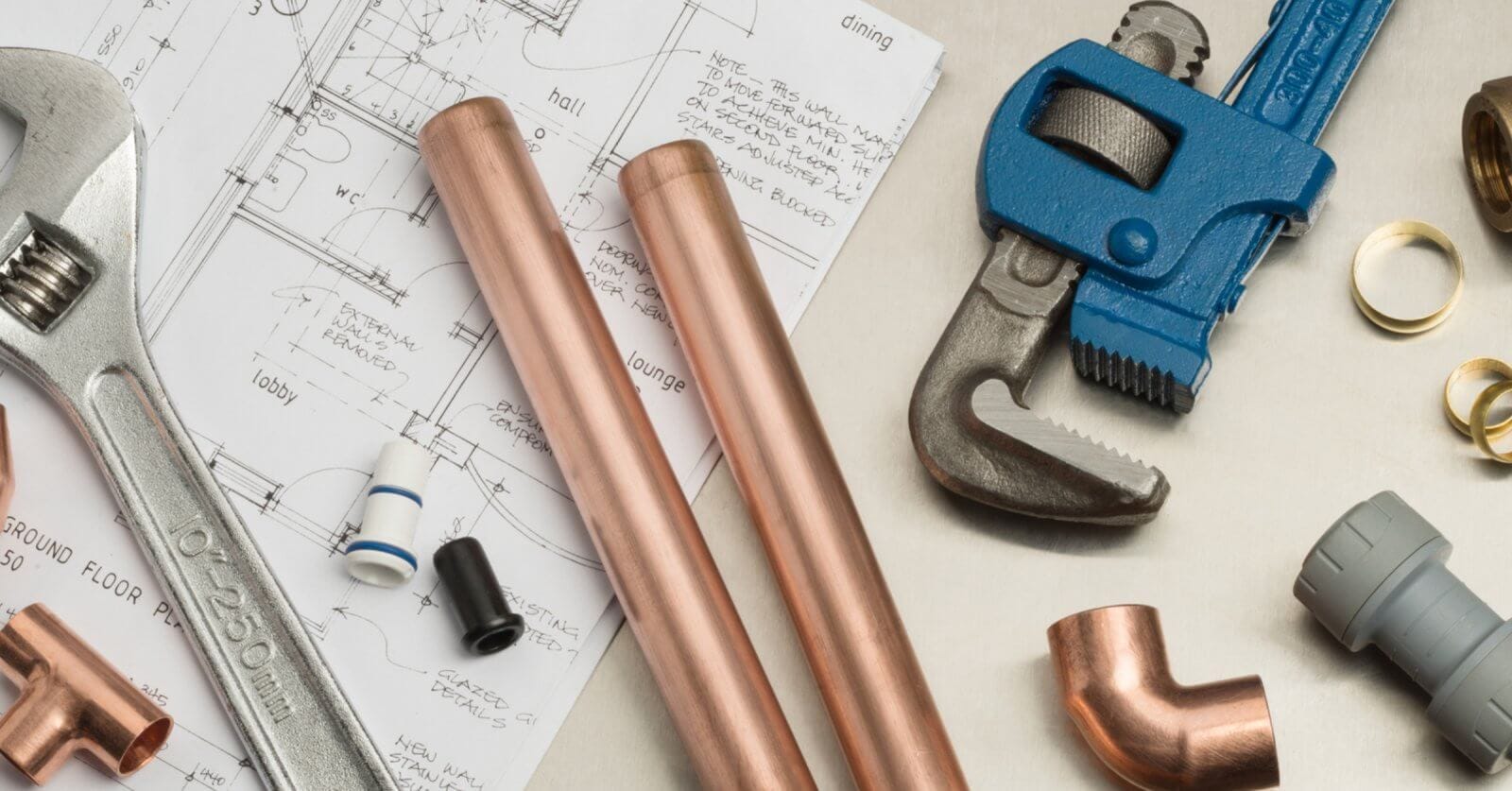 Plumbing tools and materials, ideal for plumbing services in Metro Manila, are neatly arranged on an architectural blueprint. Items include a large adjustable wrench, two copper pipes, a blue pipe wrench, copper fittings, and various pipe components set against detailed floor plans and elevations.