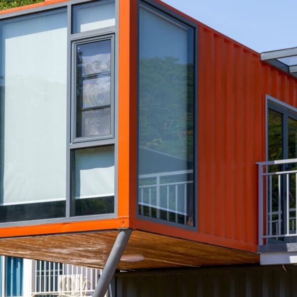 A modern structure in Metro Manila, crafted from an orange shipping container, is elevated on metal supports. It features large glass windows and a small overhanging roof. The surrounding area is lush with greenery and bathed in sunlight, while a railing suggests a balcony's presence.
