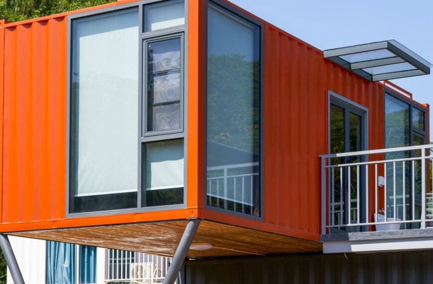 A modern structure in Metro Manila, crafted from an orange shipping container, is elevated on metal supports. It features large glass windows and a small overhanging roof. The surrounding area is lush with greenery and bathed in sunlight, while a railing suggests a balcony's presence.