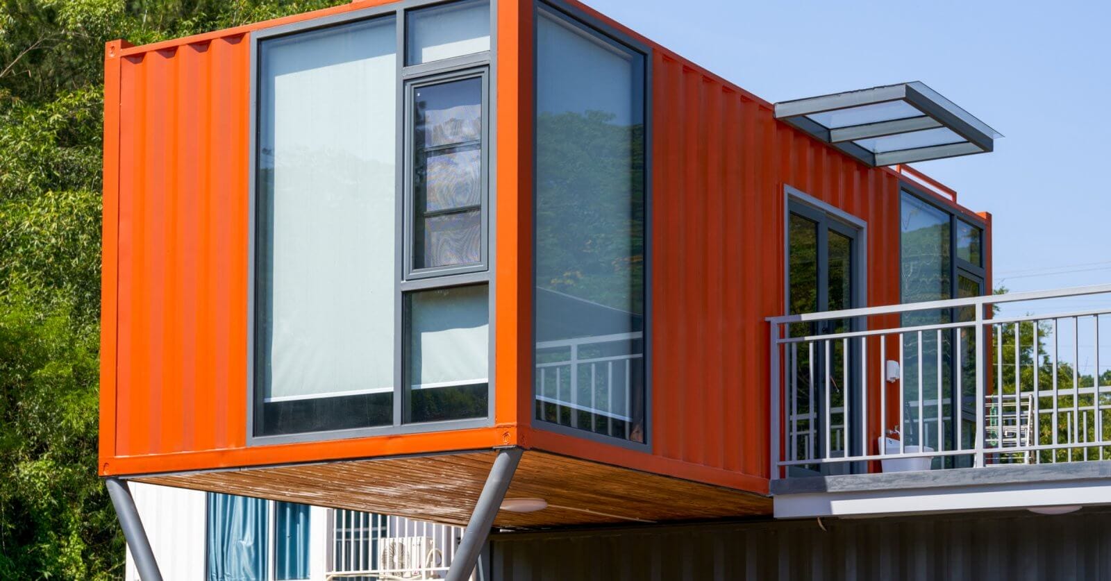 A modern structure in Metro Manila, crafted from an orange shipping container, is elevated on metal supports. It features large glass windows and a small overhanging roof. The surrounding area is lush with greenery and bathed in sunlight, while a railing suggests a balcony's presence.