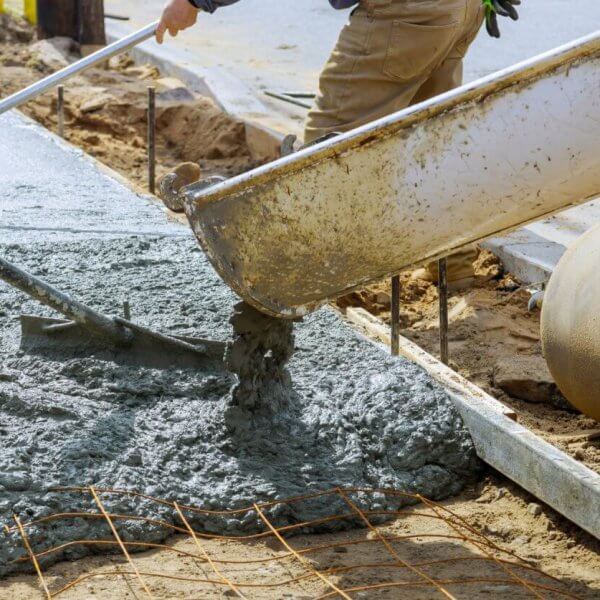 Two workers in Metro Manila pour concrete from a large chute to create a sidewalk. One uses a rake to spread the wet mix evenly. Metal reinforcing bars are visible, and a large concrete pipe lies nearby. The scene is outdoors, amid dirt and construction materials supplied by local concrete mix suppliers.