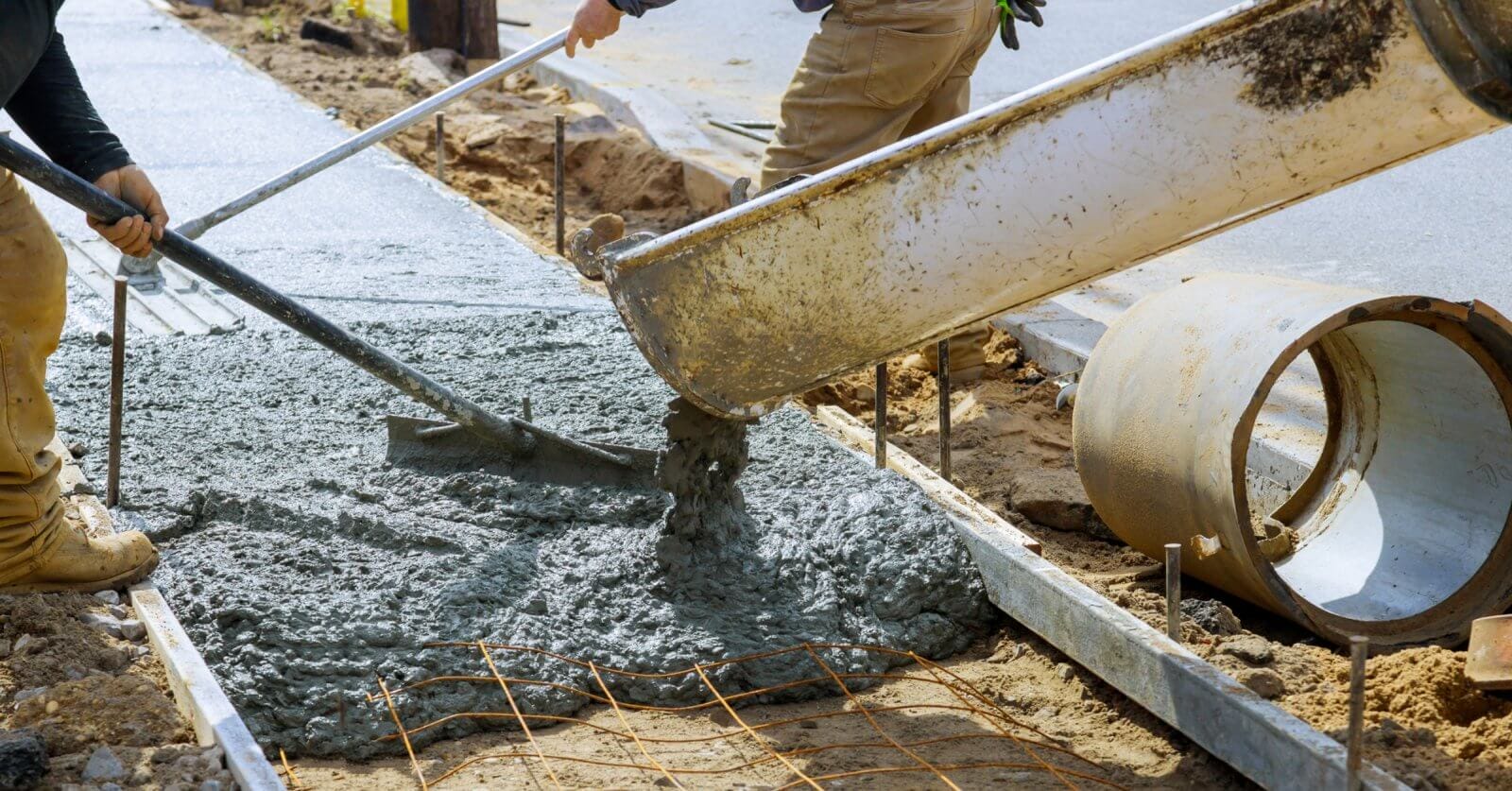 Two workers in Metro Manila pour concrete from a large chute to create a sidewalk. One uses a rake to spread the wet mix evenly. Metal reinforcing bars are visible, and a large concrete pipe lies nearby. The scene is outdoors, amid dirt and construction materials supplied by local concrete mix suppliers.