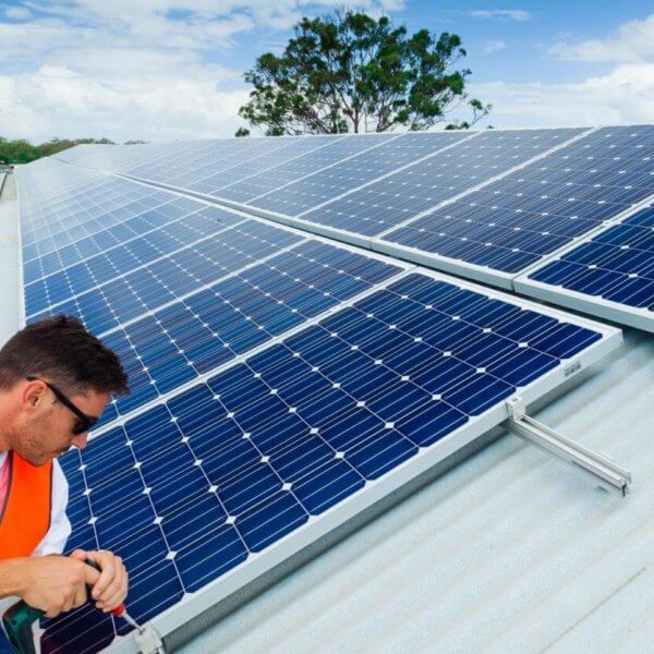 A man in an orange safety vest and sunglasses is installing solar panels on a sloped metal roof under a partly cloudy sky. Working with precision, he adjusts the panels with tools in hand. In the background, lush greenery and buildings of Metro Manila spread out, highlighting leading solar panel suppliers' impact on urban landscapes.