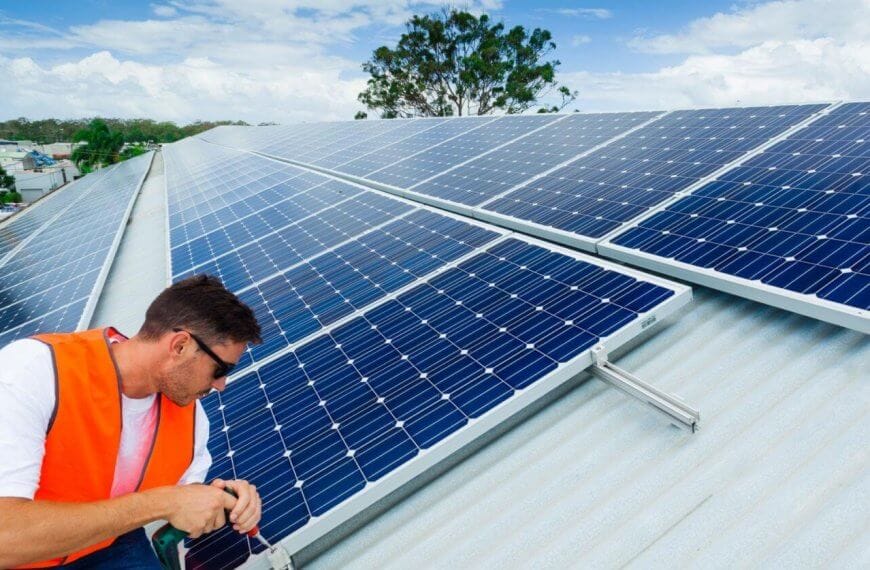 A man in an orange safety vest and sunglasses is installing solar panels on a sloped metal roof under a partly cloudy sky. Working with precision, he adjusts the panels with tools in hand. In the background, lush greenery and buildings of Metro Manila spread out, highlighting leading solar panel suppliers' impact on urban landscapes.