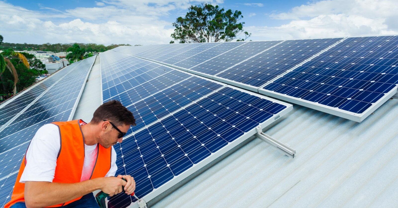 A man in an orange safety vest and sunglasses is installing solar panels on a sloped metal roof under a partly cloudy sky. Working with precision, he adjusts the panels with tools in hand. In the background, lush greenery and buildings of Metro Manila spread out, highlighting leading solar panel suppliers' impact on urban landscapes.