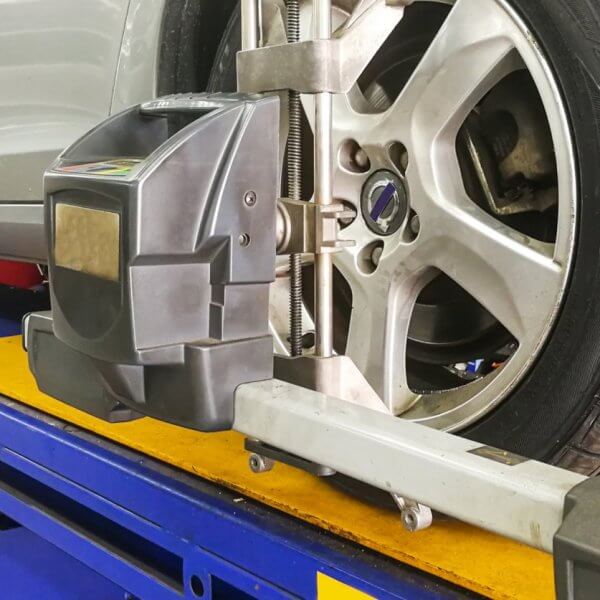 A silver car is elevated on a blue hydraulic lift in a garage in Metro Manila. A wheel alignment machine is attached to the front driver's side tire, ensuring precise alignment. The workshop environment includes tools and equipment, with clear visibility of the tire tread and alloy wheel details from one of the best centers around.