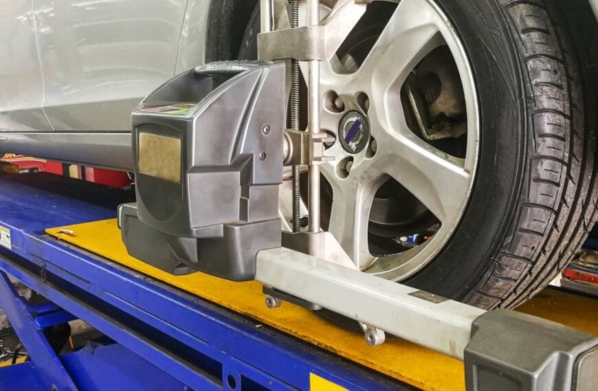 A silver car is elevated on a blue hydraulic lift in a garage in Metro Manila. A wheel alignment machine is attached to the front driver's side tire, ensuring precise alignment. The workshop environment includes tools and equipment, with clear visibility of the tire tread and alloy wheel details from one of the best centers around.