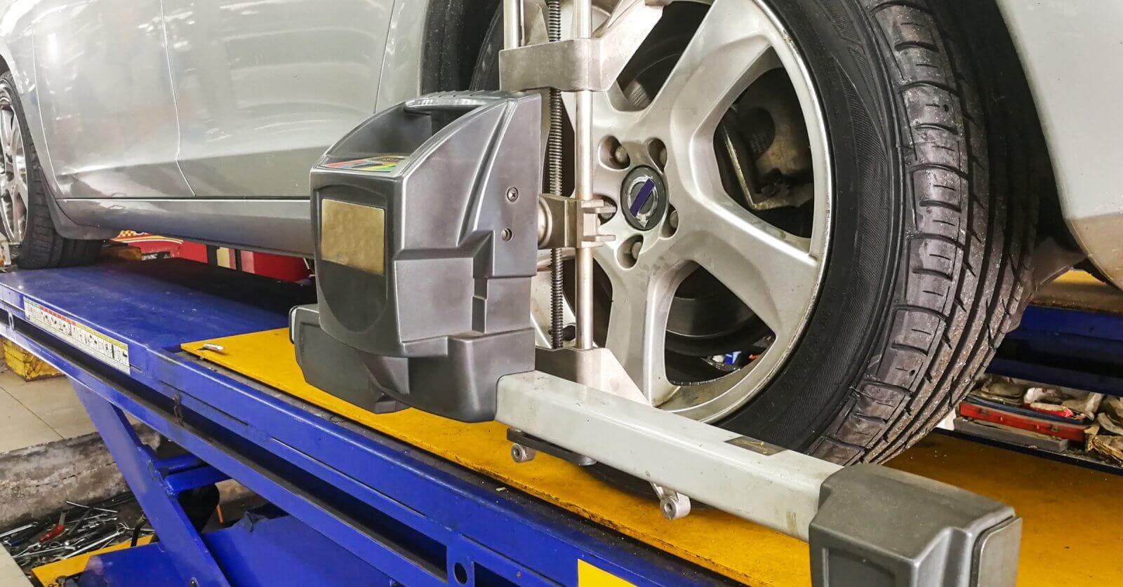 A silver car is elevated on a blue hydraulic lift in a garage in Metro Manila. A wheel alignment machine is attached to the front driver's side tire, ensuring precise alignment. The workshop environment includes tools and equipment, with clear visibility of the tire tread and alloy wheel details from one of the best centers around.
