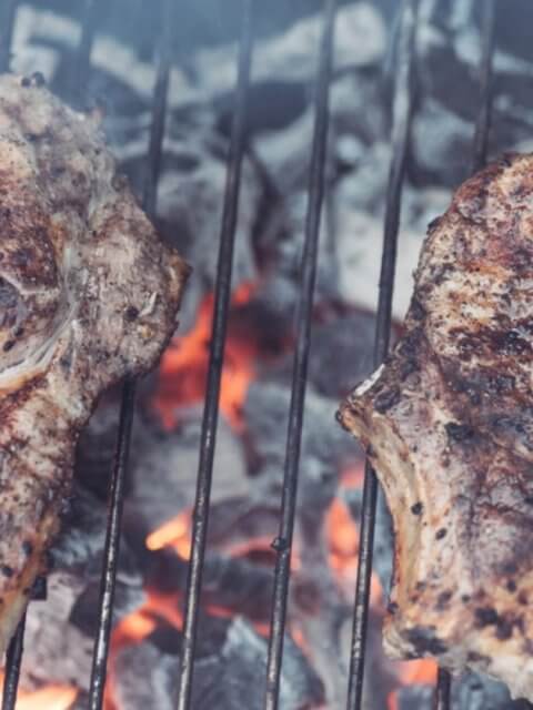 Two seasoned steaks grilling over hot charcoal, with flames and smoke visible between the grill grates, evoke a tantalizing aroma reminiscent of top Binan restaurants.