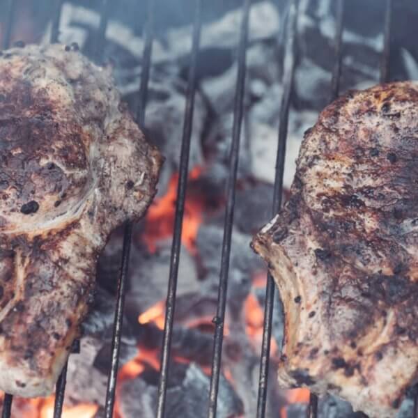Two seasoned steaks grilling over hot charcoal, with flames and smoke visible between the grill grates, evoke a tantalizing aroma reminiscent of top Binan restaurants.
