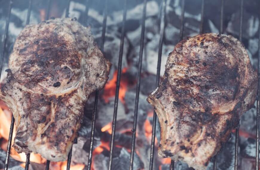 Two seasoned steaks grilling over hot charcoal, with flames and smoke visible between the grill grates, evoke a tantalizing aroma reminiscent of top Binan restaurants.