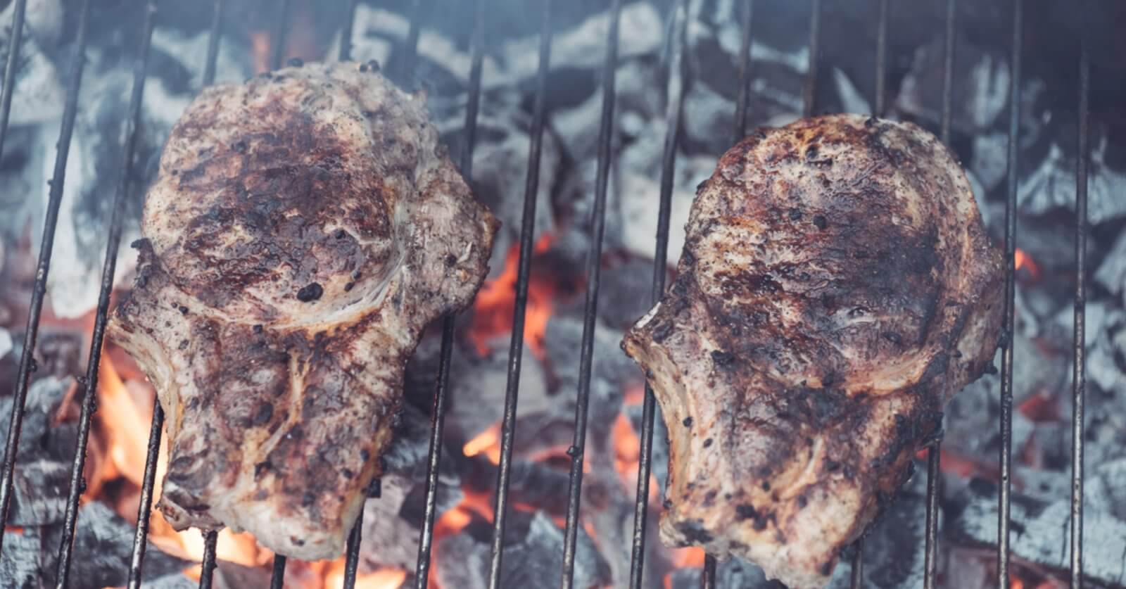 Two seasoned steaks grilling over hot charcoal, with flames and smoke visible between the grill grates, evoke a tantalizing aroma reminiscent of top Binan restaurants.