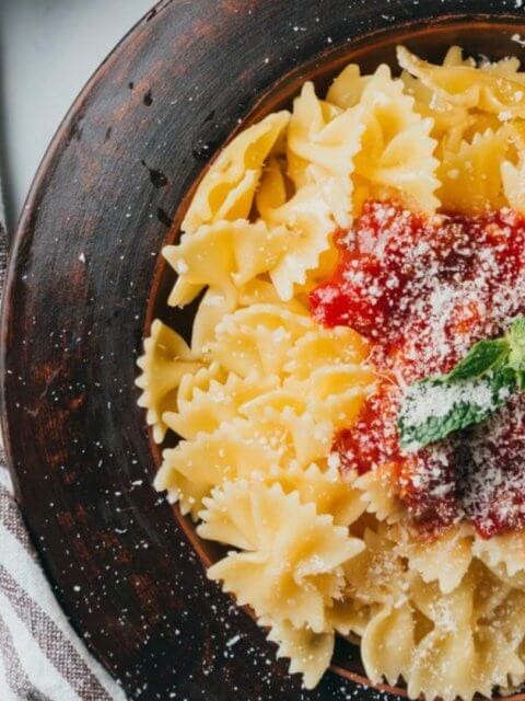 A bowl of farfalle pasta with tomato sauce and mint garnish sits elegantly next to a fork and spoon on a striped cloth, reminiscent of the culinary delights found in Ayala Malls' top restaurants.