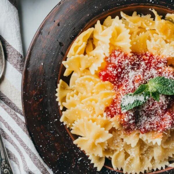A bowl of farfalle pasta with tomato sauce and mint garnish sits elegantly next to a fork and spoon on a striped cloth, reminiscent of the culinary delights found in Ayala Malls' top restaurants.