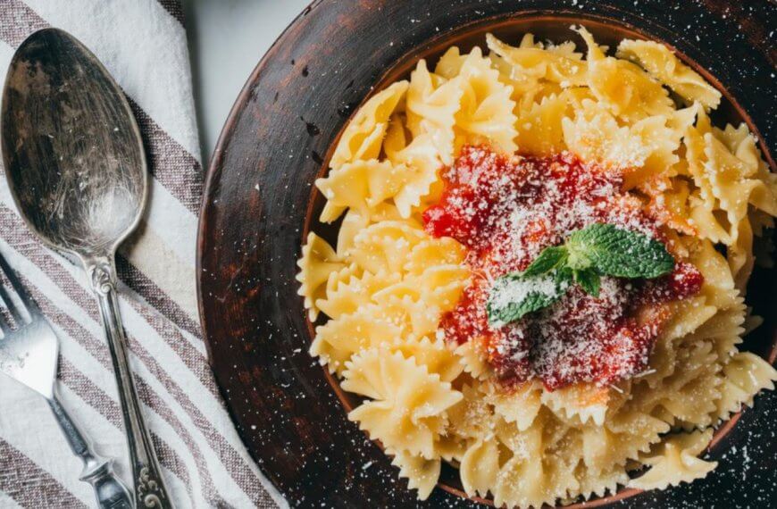A bowl of farfalle pasta with tomato sauce and mint garnish sits elegantly next to a fork and spoon on a striped cloth, reminiscent of the culinary delights found in Ayala Malls' top restaurants.