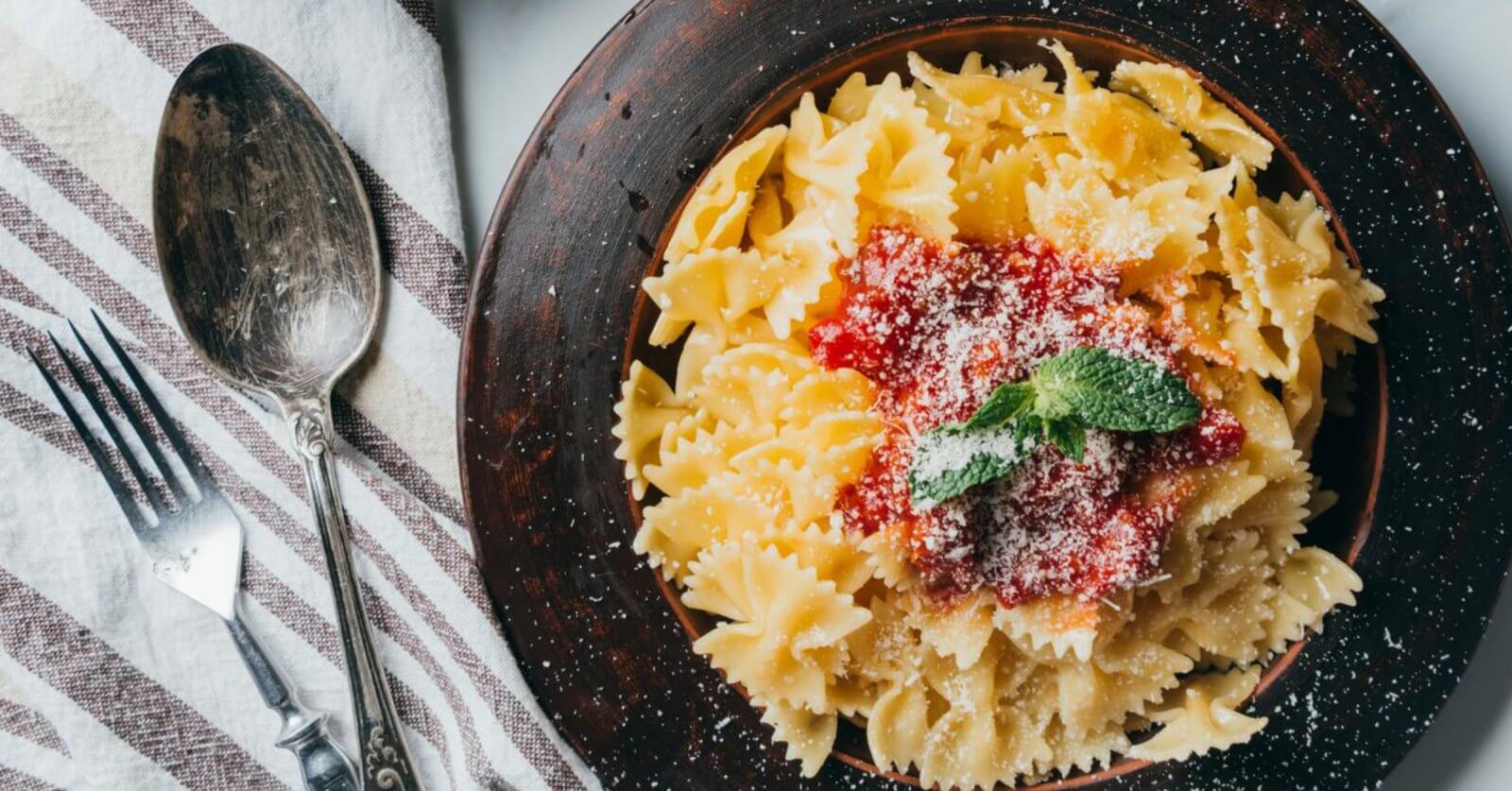 A bowl of farfalle pasta with tomato sauce and mint garnish sits elegantly next to a fork and spoon on a striped cloth, reminiscent of the culinary delights found in Ayala Malls' top restaurants.