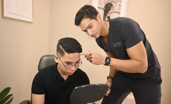 Two men in black shirts, one with a mirror and the other pointing at his head, in a Clinique de Paris consultation room.