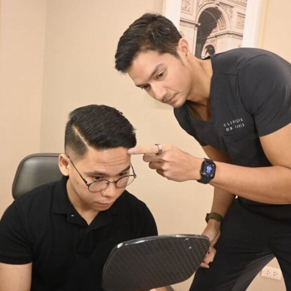 Two men in black shirts, one with a mirror and the other pointing at his head, in a Clinique de Paris consultation room.