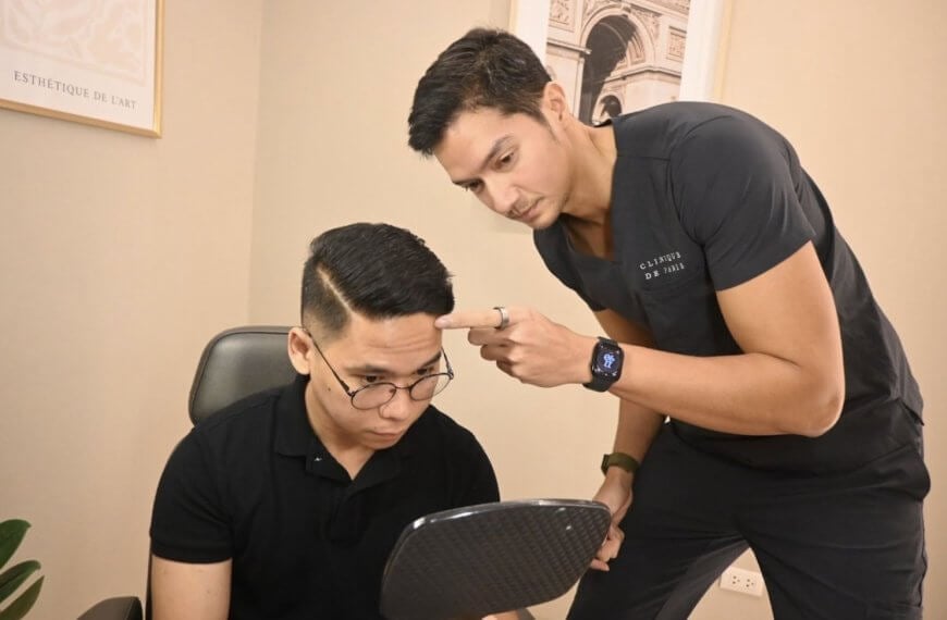 Two men in black shirts, one with a mirror and the other pointing at his head, in a Clinique de Paris consultation room.