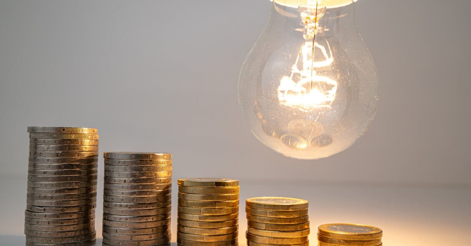 Stacked coins beneath a lit bulb, symbolizing electric consumption and the potential to save with solar.